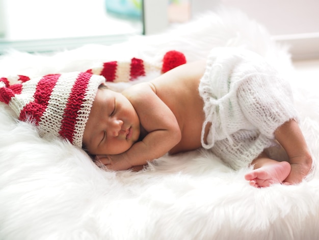 Bebé recién nacido asiático vistiendo un gorro de duende navideño tejido durmiendo sobre pelaje blanco