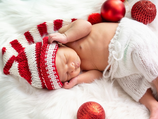 Bebé recién nacido asiático vistiendo un gorro de duende navideño tejido durmiendo sobre pelaje blanco