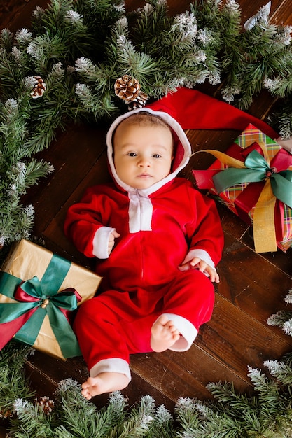 Bebé recién nacido disfrazado de Santa Claus se encuentra en medio de una corona de árboles de Navidad