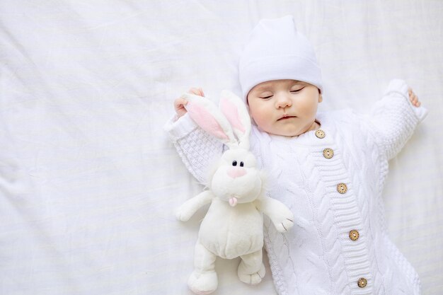 Un bebé recién nacido duerme en una cuna con un mono blanco cálido y un sombrero en una cama de algodón blanco con un juguete suave para dormir sano.