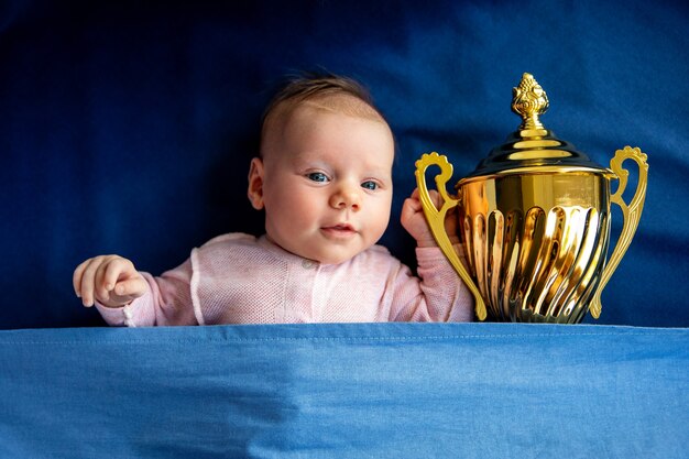 Bebé recién nacido mirando a la cámara, niña con copa de oro, concepto de victoria