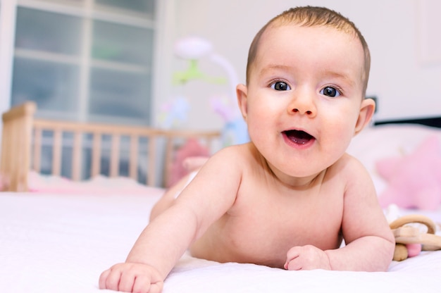 Bebé de risa feliz lindo después del baño que juega en la cama blanca con las almohadas en sitio soleado de los niños.