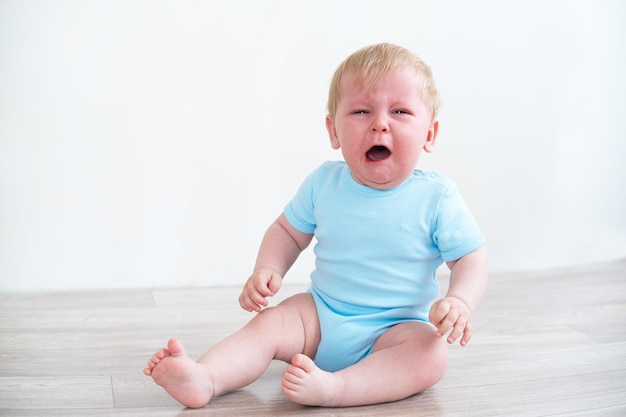 bebé rubio con camisa azul llorando sentado en el suelo en casa
