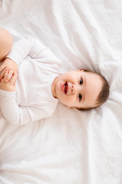 Bebé sano de 10 meses de edad con ropa blanca sonriendo sentado en ropa de cama blanca en la cama, espacio para texto