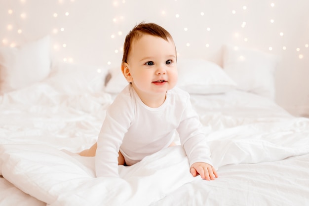 Bebé sano de 10 meses de edad con ropa blanca sonriendo sentado en ropa de cama blanca en la cama, espacio para texto