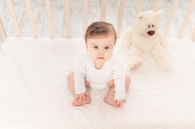 Bebé de seis meses sentado en una cuna con un mono blanco con juguetes Ositos de peluche