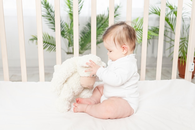Bebé de seis meses sentado en una cuna con un mono blanco con juguetes Ositos de peluche