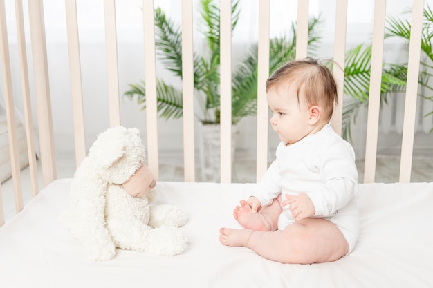 Bebé de seis meses sentado en una cuna con un mono blanco con juguetes Ositos de peluche