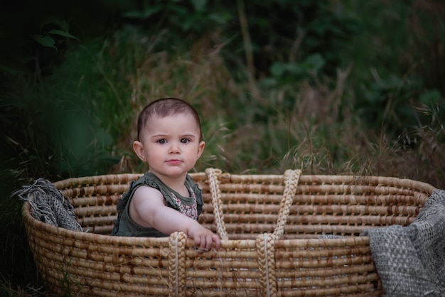El bebé se sienta en una cuna de mimbre de paja en un parque verde. Niña en vestido lindo sonriendo. Retrato.