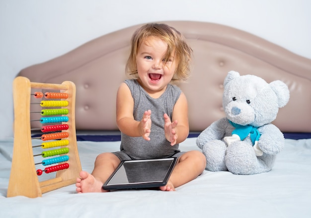 El bebé se sienta con juguetes y la tableta en la cama y felizmente aplaude