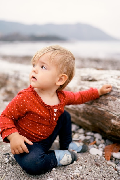 El bebé se sienta en una playa de guijarros cerca de una vieja madera flotante y gira la cabeza hacia un lado