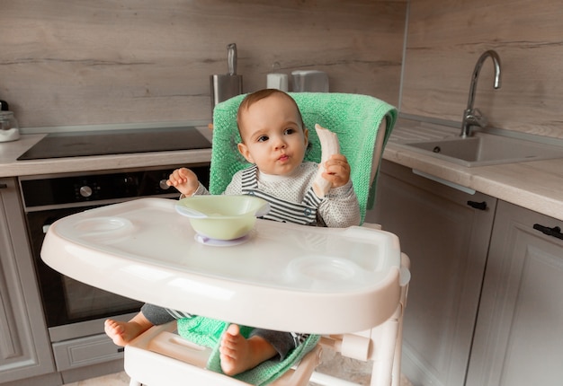 El bebé se sienta en una silla alta en la cocina y come un plátano.