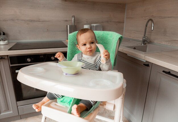 El bebé se sienta en una silla alta en la cocina y come un plátano.