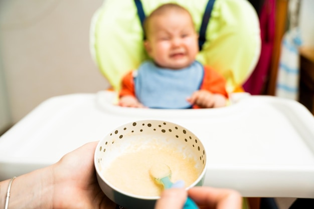 Foto el bebé en la silla alta no quiere comer la comida que la madre le da con una cuchara traviesa y llorando