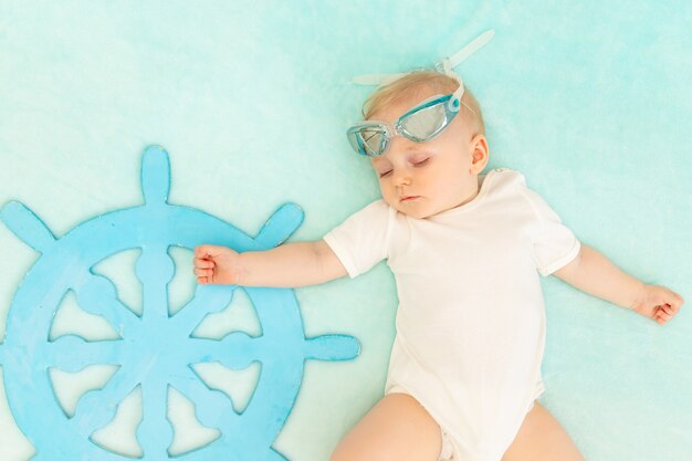 Un bebé sobre un fondo azul duerme con el volante de un barco y gafas de natación