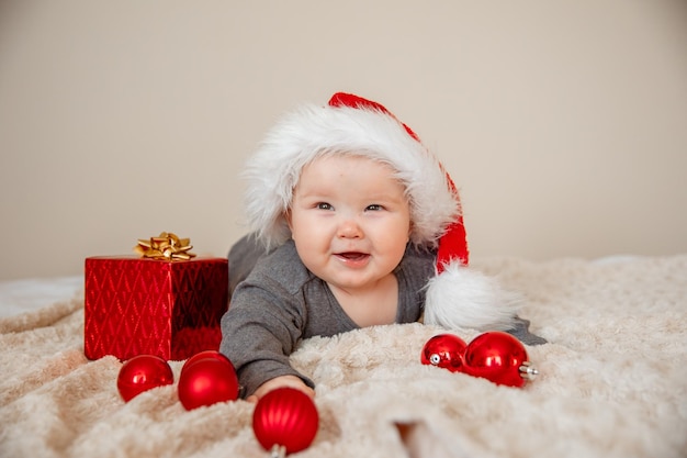 Un bebé con un sombrero de Papá Noel está acostado en la cama jugando con bolas de Navidad