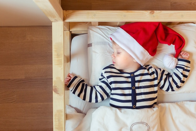 Bebê sonolento com chapéu de Papai Noel vermelho na cama