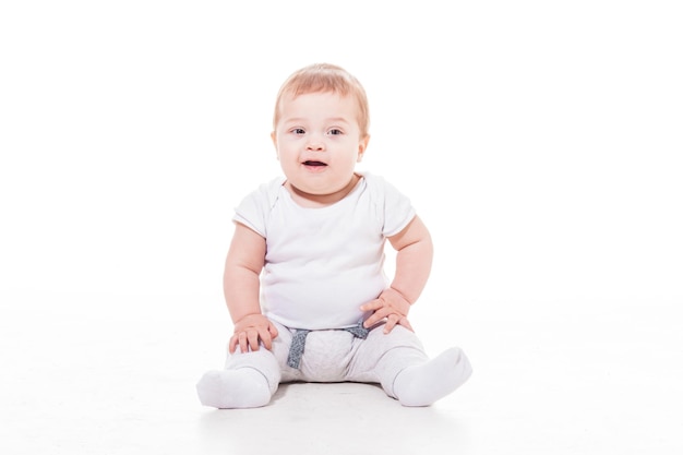 Bebé sonriente feliz, vestido con un paño blanco está sentado en el suelo sobre fondo blanco.