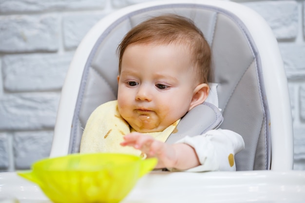 Un bebé sonriente de nueve meses se sienta en una mesa blanca en una trona y come con una cuchara de un tazón. Mamá alimenta al bebé con una cuchara. Fondo borroso. Alimentos saludables para niños. Alimentos para niños.