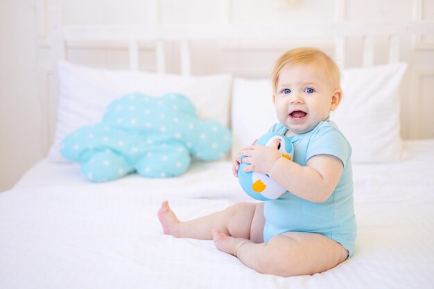 Un bebé sonriente o riendo aprende a gatear en la cama en casa con ropa azul, el bebé juega con un juguete de sonajero de plástico para acostarse feliz infancia y familia