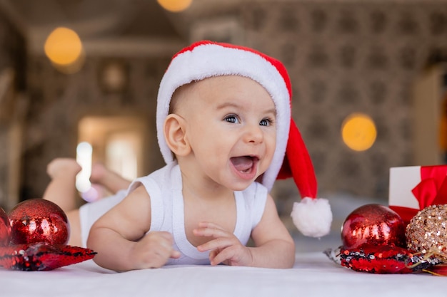 Bebê sorridente bonito em uma roupa branca e chapéu de Papai Noel está deitado de bruços em um lençol branco
