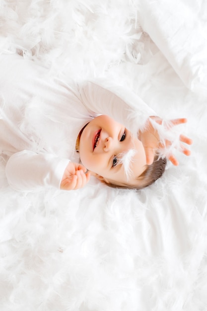 bebê sorridente com roupas brancas deitado de costas na cama em um lençol branco e brincando com penas