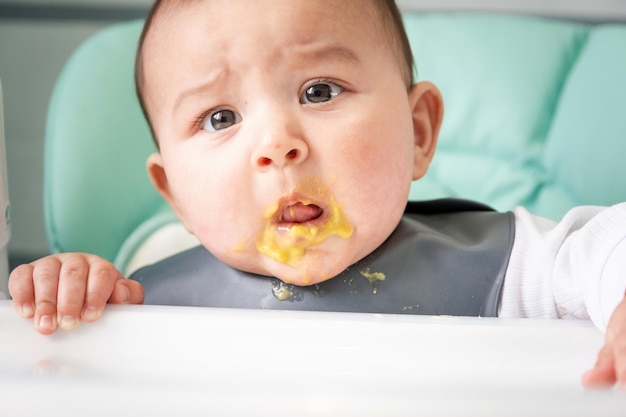 Un bebé sucio y sorprendido manchado de puré de verduras en la mesa de alimentación Introducción de alimentos complementarios no les gusta el sabor de la comida nueva Retrato de cierre