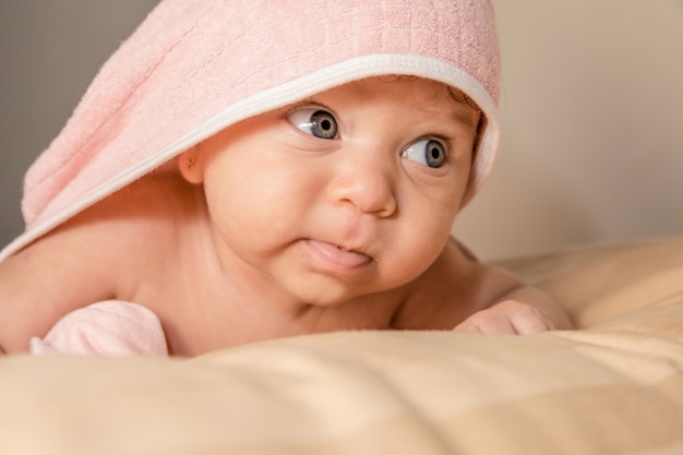 Bebê surpreendido com grandes olhos azuis e boca aberta deitada na barriga sob a toalha rosa após banho