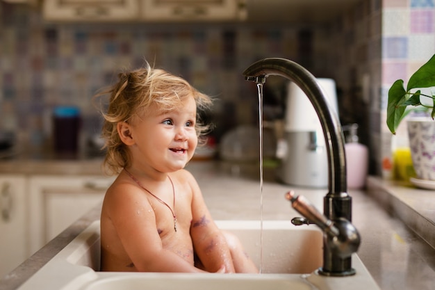 Bebé tomando baño en el fregadero de la cocina. niño jugando con agua en cocina soleada con ventana.