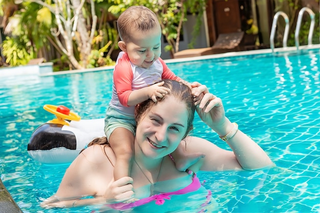 Bebé en traje de baño sentarse sobre los hombros de la madre y juega con el pelo en la cabeza tira del pelo en la piscina