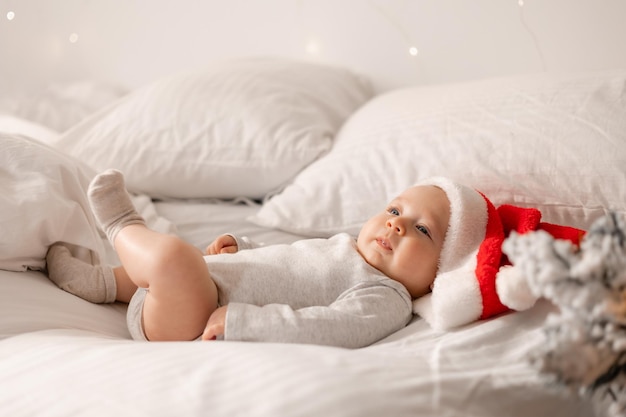 El bebé con un traje blanco y un gorro de Papá Noel está acostado en la cama junto al árbol de Navidad. encantador niño de ojos azules. vacaciones de invierno, momentos divertidos, navidad infantil. espacio para texto. foto de alta calidad