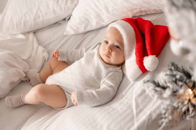 El bebé con un traje blanco y un gorro de Papá Noel está acostado en la cama junto al árbol de Navidad. encantador niño de ojos azules. vacaciones de invierno, momentos divertidos, navidad infantil. espacio para texto. foto de alta calidad