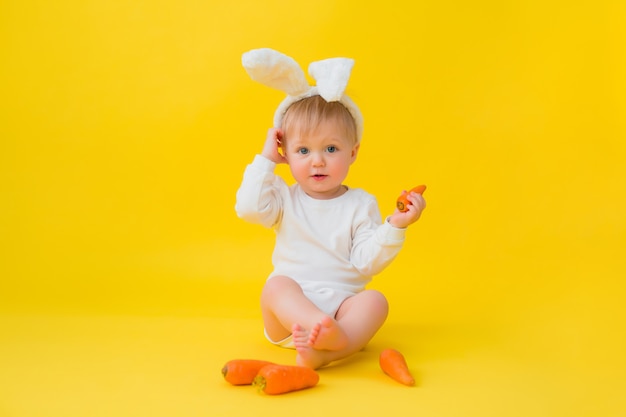 Bebé en un traje blanco con orejas de conejo en la cabeza come zanahorias, se sienta sobre un fondo amarillo con verduras. bebé en forma de conejito de pascua, espacio para texto