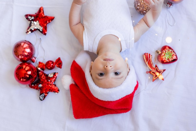 El bebé con un traje blanco y un sombrero de Papá Noel está acostado de espaldas sobre una sábana blanca rodeada de juguetes rojos de árboles de Navidad. invierno, año nuevo. espacio para texto. foto de alta calidad