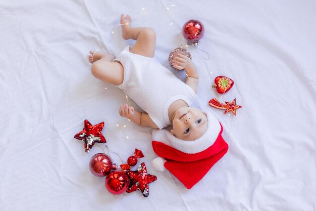 El bebé con un traje blanco y un sombrero de Papá Noel está acostado de espaldas sobre una sábana blanca rodeada de juguetes rojos de árboles de Navidad. invierno, año nuevo. espacio para texto. foto de alta calidad