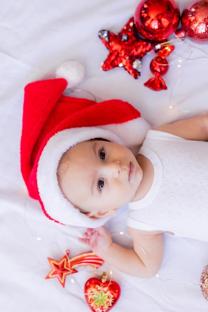 El bebé con un traje blanco y un sombrero de Papá Noel está acostado de espaldas sobre una sábana blanca rodeada de juguetes rojos de árboles de Navidad. invierno, año nuevo. espacio para texto. foto de alta calidad