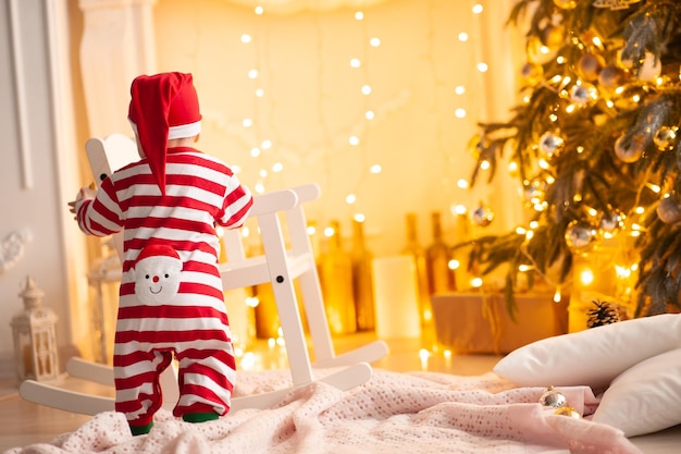 Bebé con traje de Papá Noel parado en el árbol de Navidad en la habitación. Vista trasera. Temporada de vacaciones.