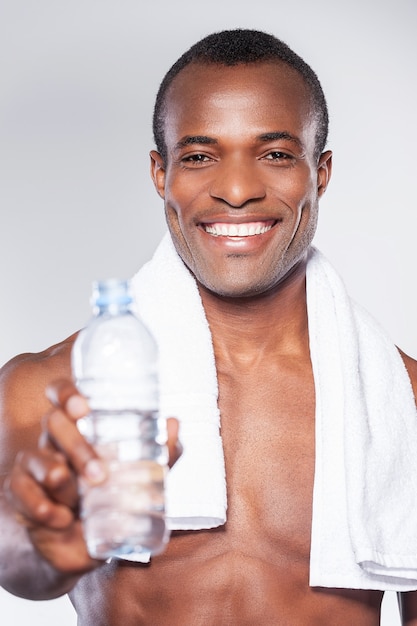 Bebe um pouco de água! jovem africano musculoso esticando uma garrafa com água na mão e sorrindo