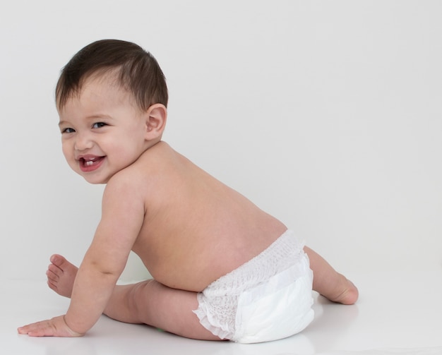 El bebé usa un pañal blanco, se sienta girando la espalda y sonríe
