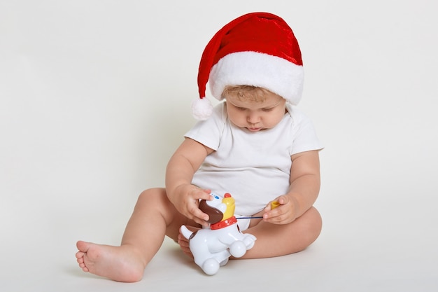 Bebê veste chapéu de Papai Noel posando isolado sobre uma parede branca enquanto está sentado com os pés descalços no chão e brincando com cachorro de plástico, criança olhando para o brinquedo com interesse.
