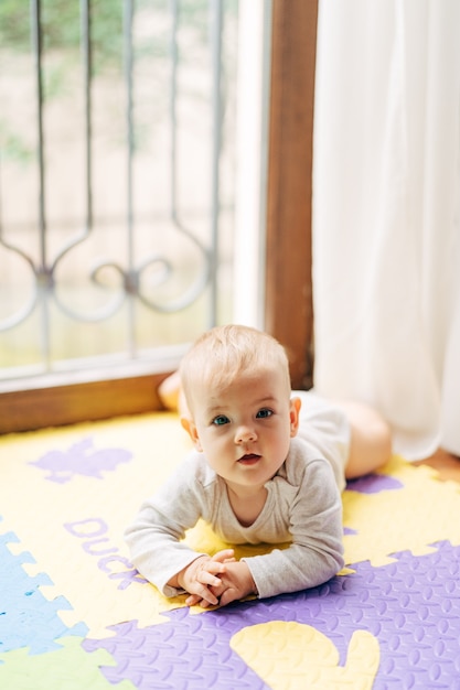 El bebé yace boca abajo sobre una alfombra de colores en el suelo con el telón de fondo de una ventana