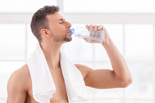 Beber agua después del entrenamiento. Hombre musculoso joven guapo con una toalla sobre los hombros bebiendo agua y manteniendo los ojos cerrados