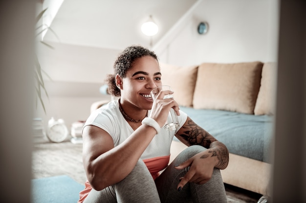 Beber água limpa. mulher sorridente e positiva de cabelos escuros bebendo água de um copo alto após um treino ativo