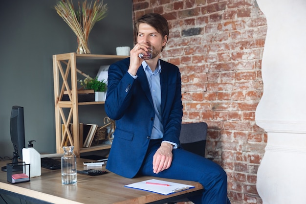 Beber agua sentada sobre la mesa. El joven, gerente, vuelve a trabajar en su oficina después de la cuarentena, se siente feliz e inspirado. Volviendo a la vida normal. Negocios, finanzas, concepto de emociones.