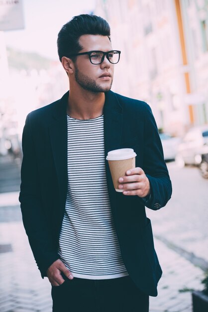 Beber café sobre la marcha. Apuesto joven en ropa casual elegante caminando por la calle mientras sostiene la taza de café