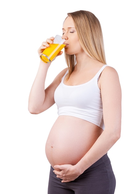 Beber jugo fresco. Hermosa mujer embarazada bebiendo jugo de naranja mientras está de pie aislado en blanco