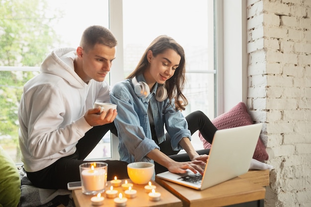 Beber té, ver cine, lucir feliz. Bloqueo de cuarentena, concepto de estancia en casa: joven hermosa pareja caucásica disfrutando de un nuevo estilo de vida durante el coronavirus. Felicidad, unión, salud.