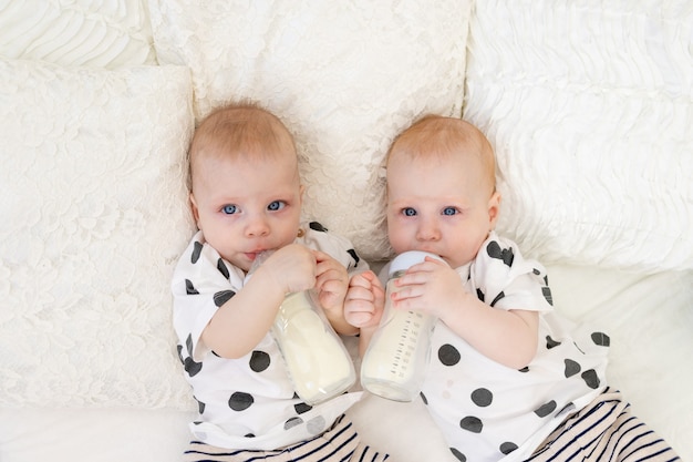 Los bebés gemelos se acuestan en la cama en pijama y beben leche de biberones