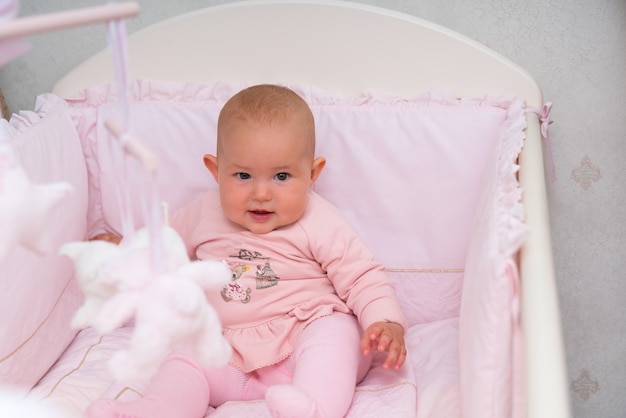Bebezinha linda sentada em um berço rosa olhando curiosamente para a câmera em uma visão de perto