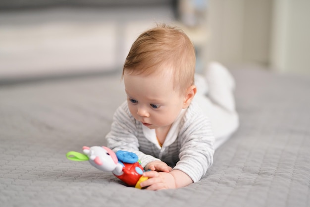 Bebezinho bonitinho segurando o brinquedo sorrindo alegremente para a câmera no quarto branco ensolarado e brilhante